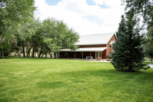 A spacious green lawn in front of a red house surrounded by trees under a partly cloudy sky.